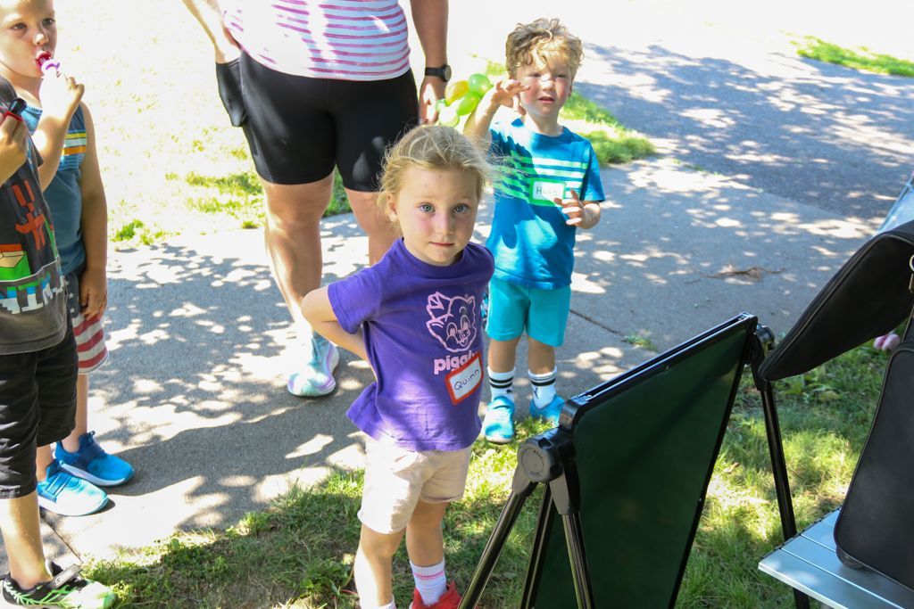 Children in a sunny outdoor setting; one child wearing a purple shirt, others near adults.