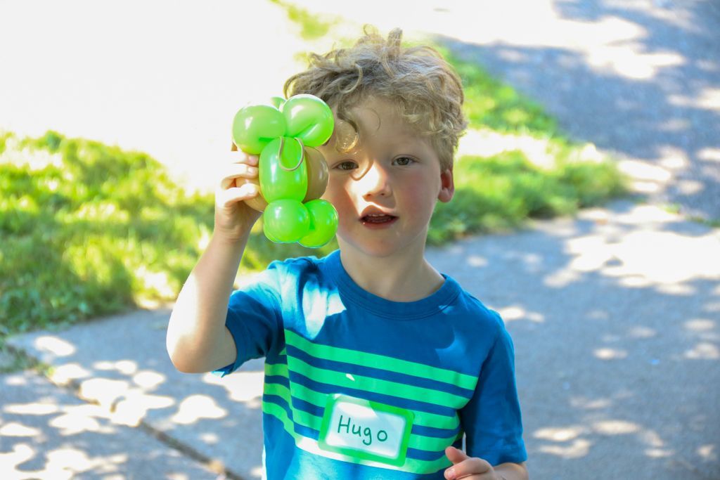 Boy with curly hair holds a green balloon animal outside. He wears a blue striped shirt with 
