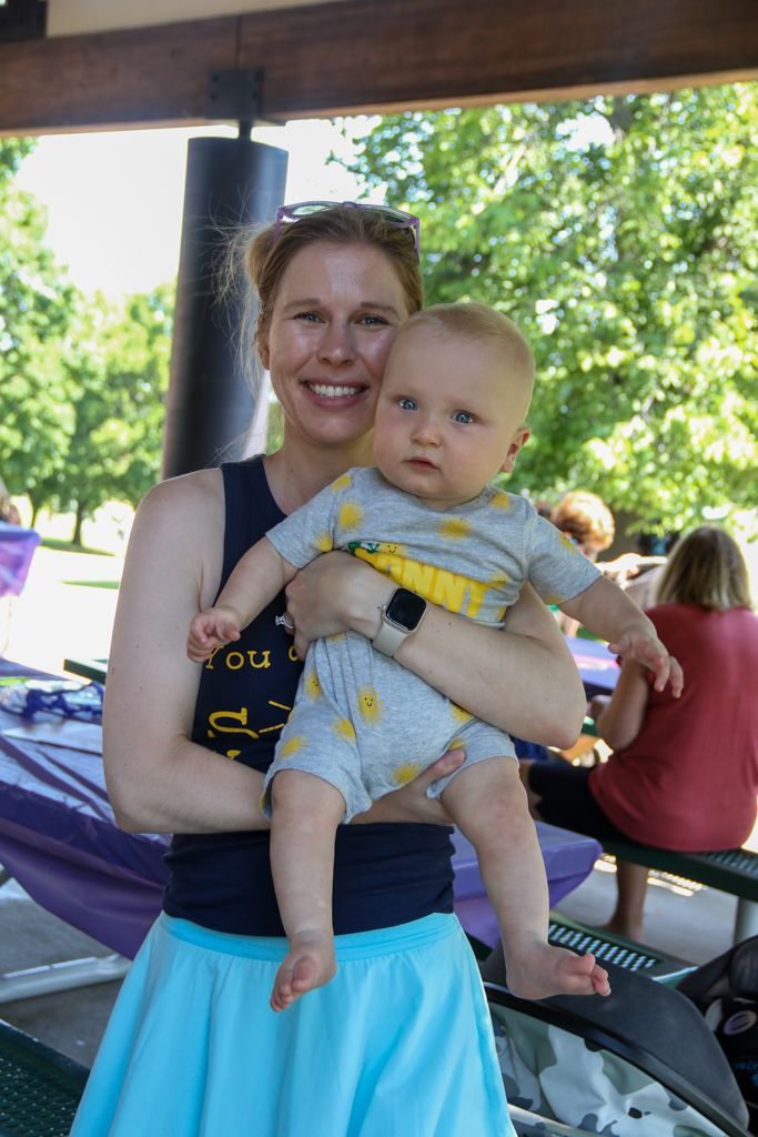 Woman holding a baby outdoors. The woman is smiling, baby looking forward. Park setting, sunny.