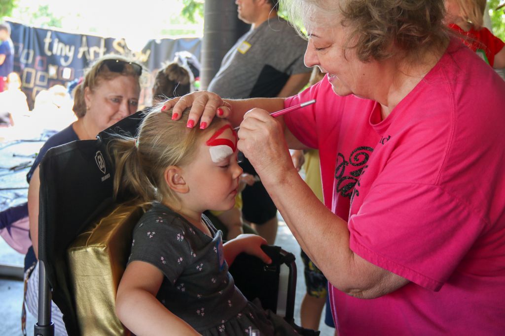 Woman paints a design on a child's face outdoors.