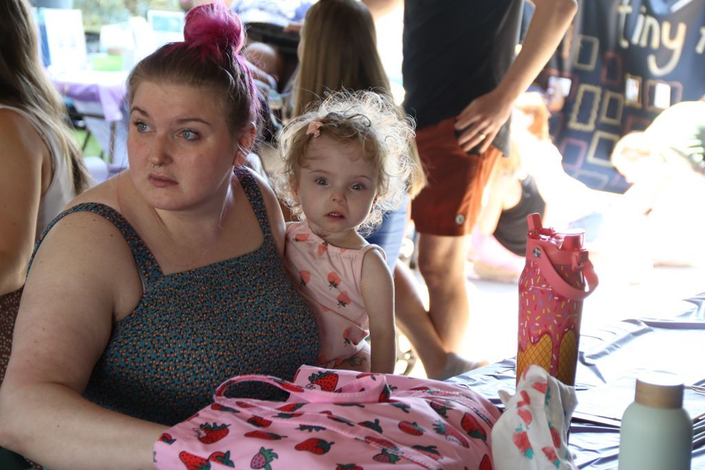 Woman with pink hair holds a child wearing a strawberry dress at an outdoor event.