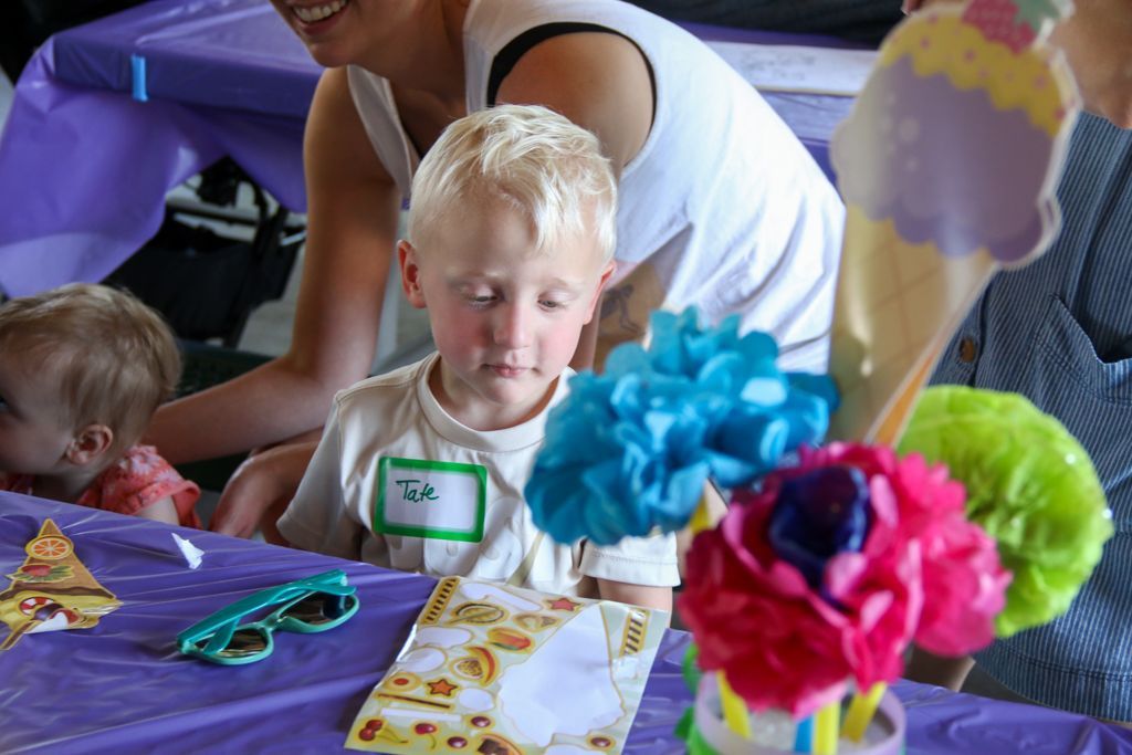 Blond child at purple table with craft supplies, looking down. Woman and another child in background.