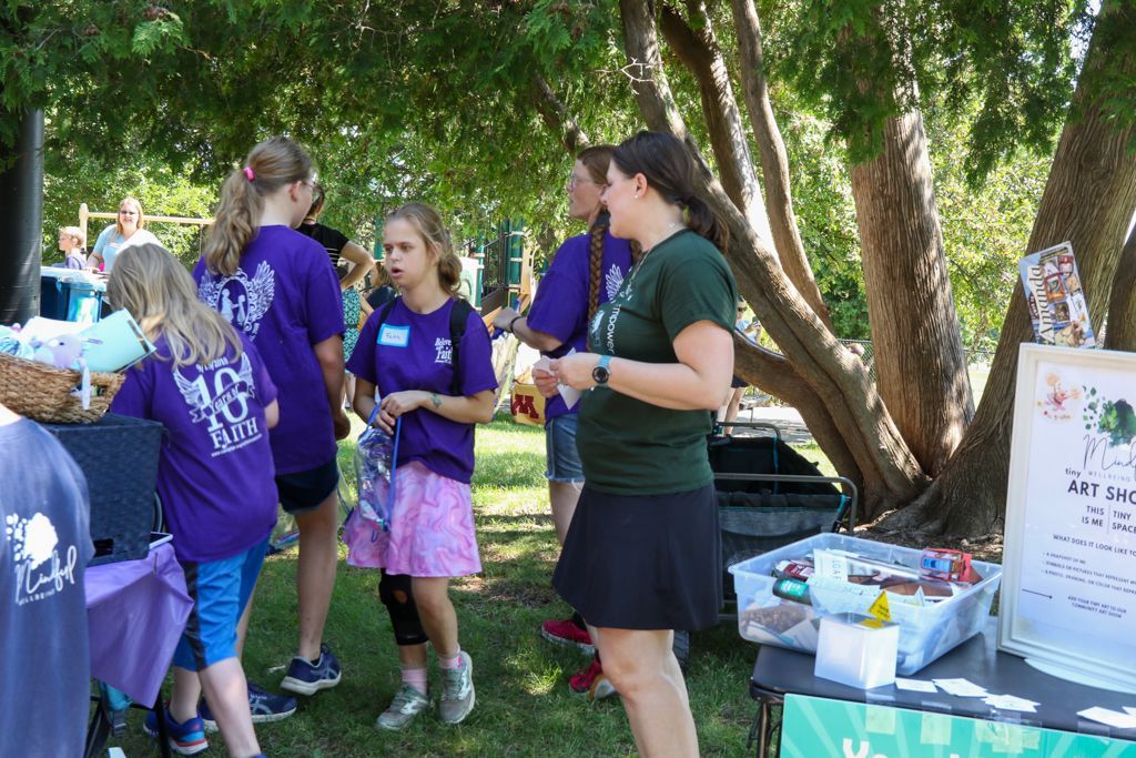People at an outdoor event with a table under a tree. Children in purple shirts and an adult are present.
