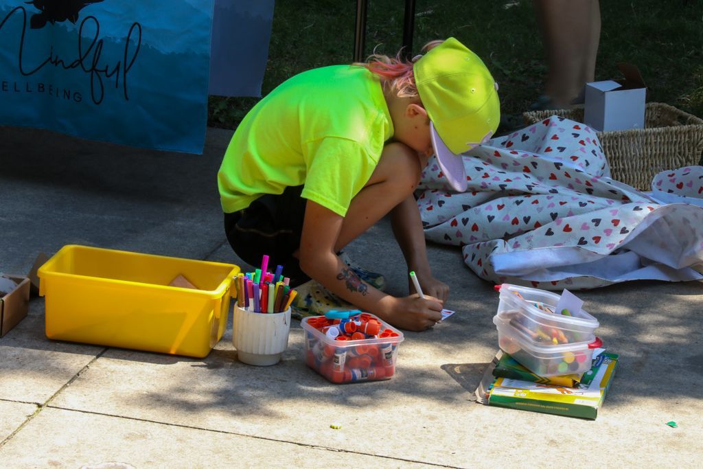 Person in neon shirt and cap, squatting, drawing on pavement. Art supplies and boxes nearby.
