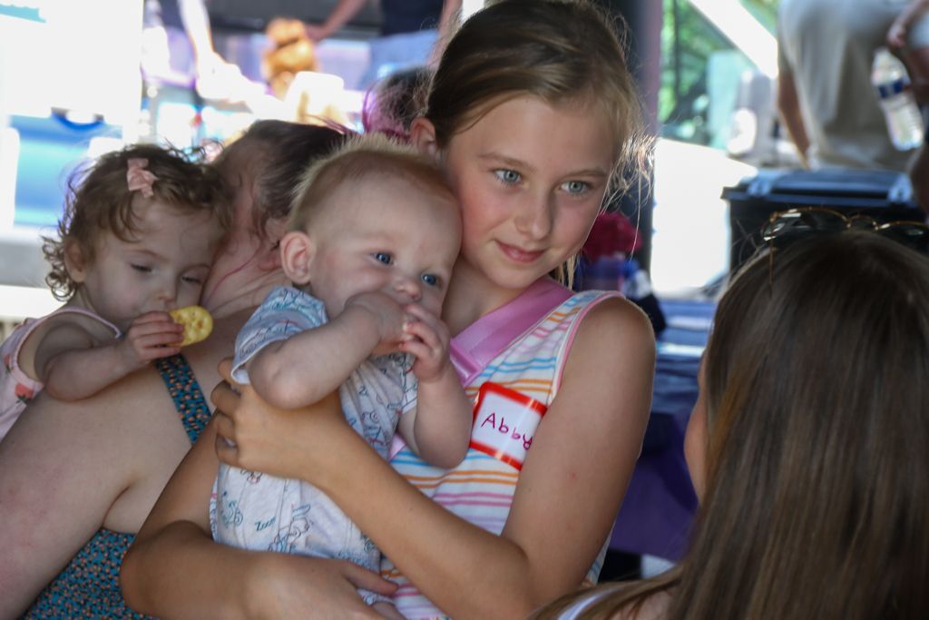Girl holding baby, looking at another person. Other children in background, outdoors.