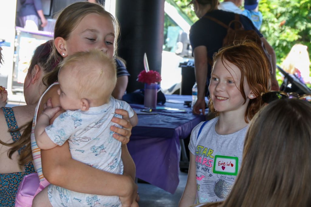 Girl holding a baby, talking to a smiling girl. Outdoors at a gathering with tables and people.
