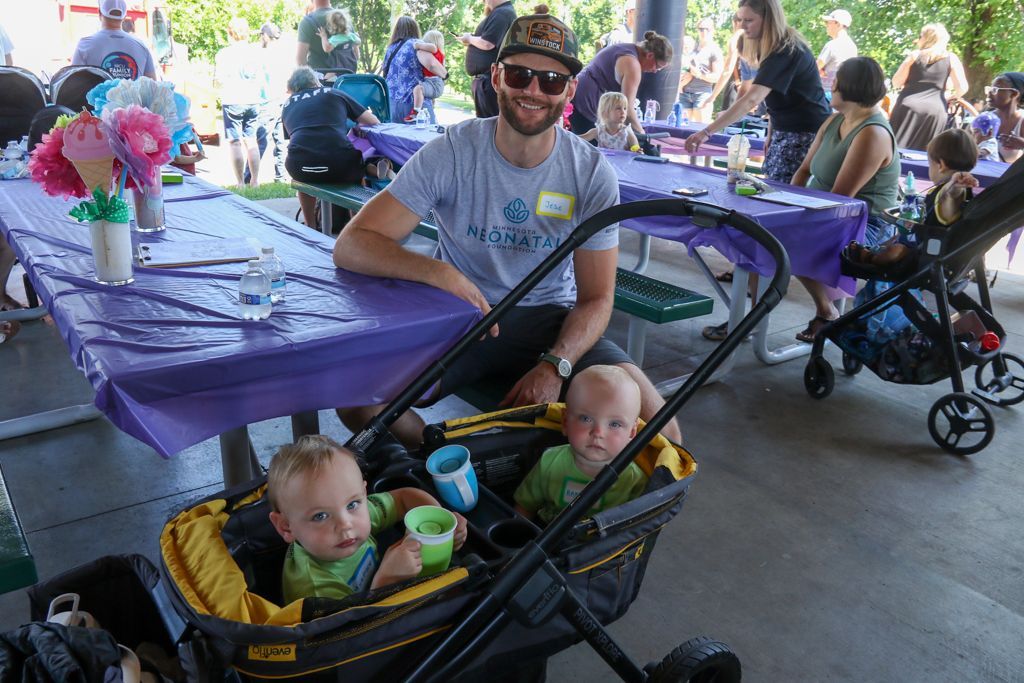 Man smiling, sitting near two babies in a stroller, at an outdoor event with purple tablecloths.