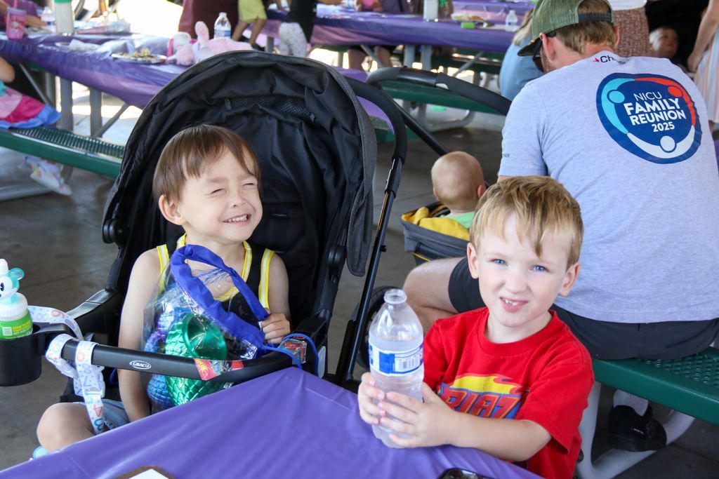 Two young children smiling at the camera near a picnic table. One in a stroller, the other holding a water bottle.