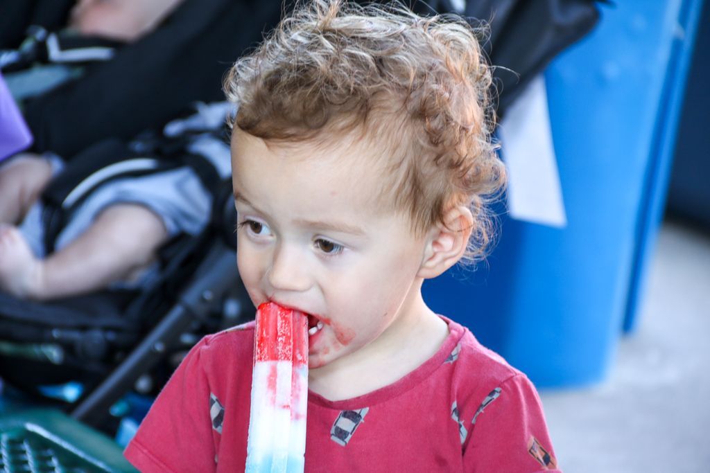 Child eating a red, white, and blue popsicle. Curly hair, red shirt, stroller in background.