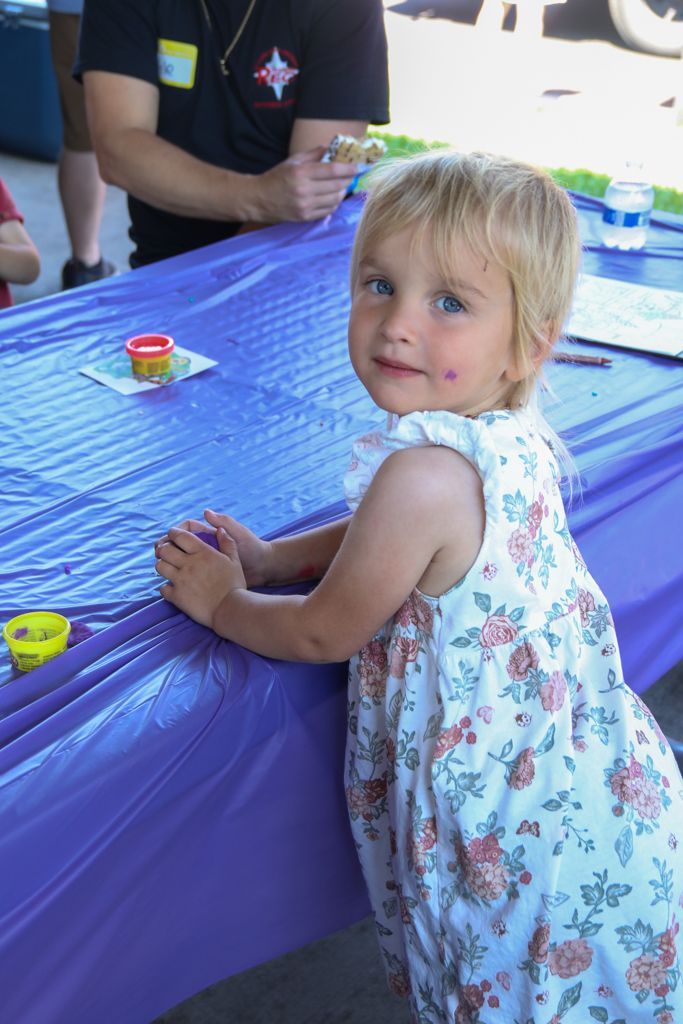 Young child in floral dress at a purple table, looking at the camera with a playful expression.