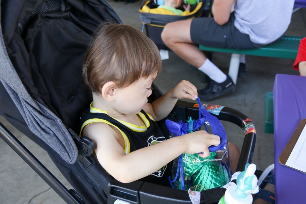 Child in stroller, looking at a blue bag. Black and yellow tank top. Someone seated in the background.