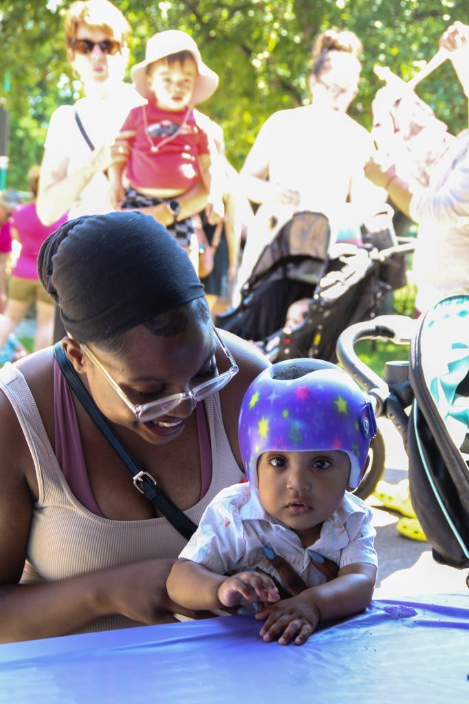 Woman holding a baby wearing a purple helmet. People and strollers in the park are in the background.