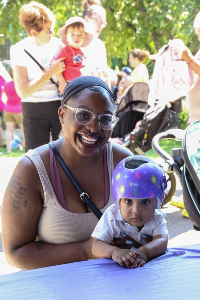 Woman smiling with baby in a helmet at an outdoor event. Other people and strollers in the background.