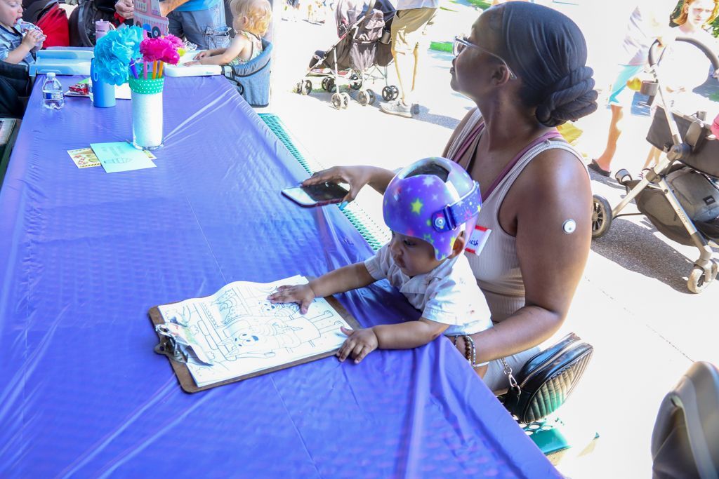 Woman holding baby wearing a helmet at a table, both looking at a clipboard. Outdoors.