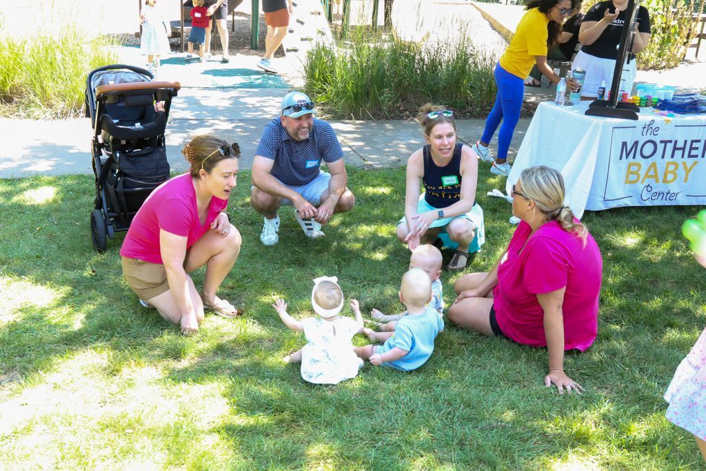 People at an outdoor event with baby dolls on the grass. A table has a sign that reads 