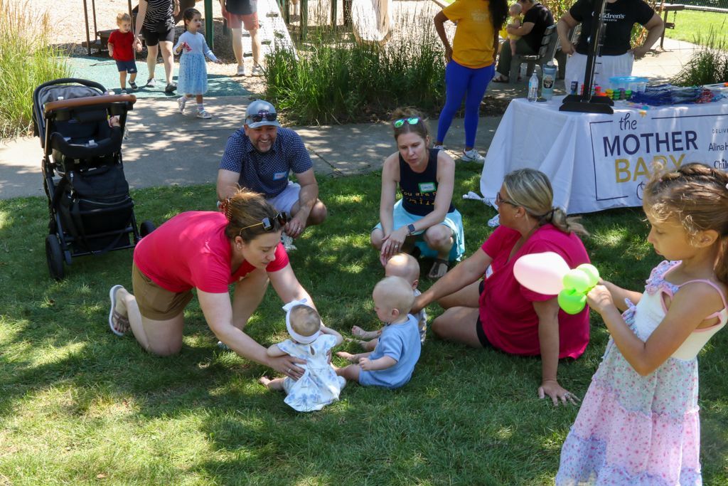 People and babies at outdoor event with a table that reads 