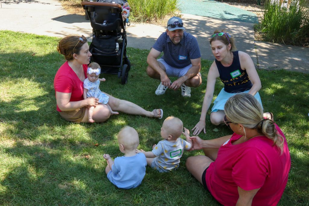 Adults and babies seated in a circle on grass. People are talking, babies are playing. Sunny day.