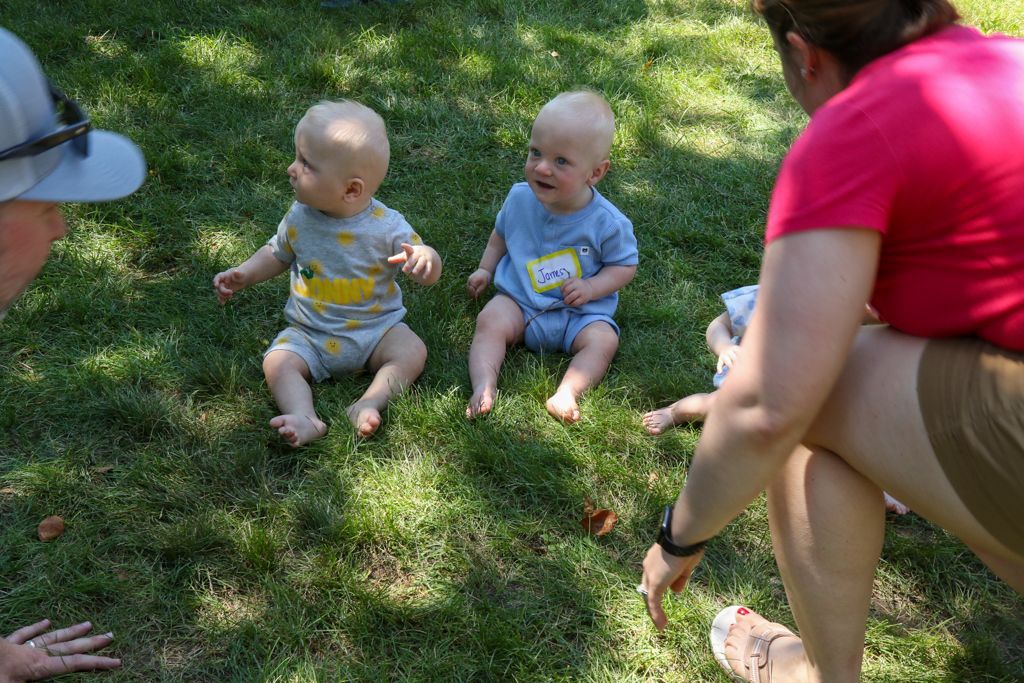 Two babies sit on grass, facing away from adults. Adults point toward babies.