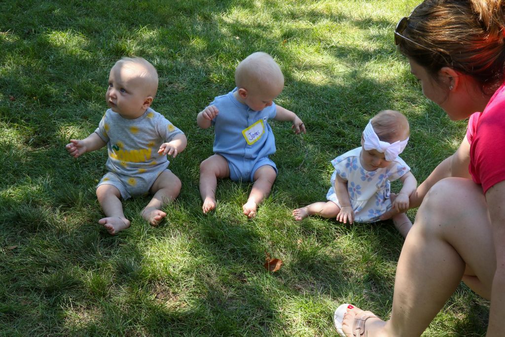Three babies sit on grass, observed by an adult.