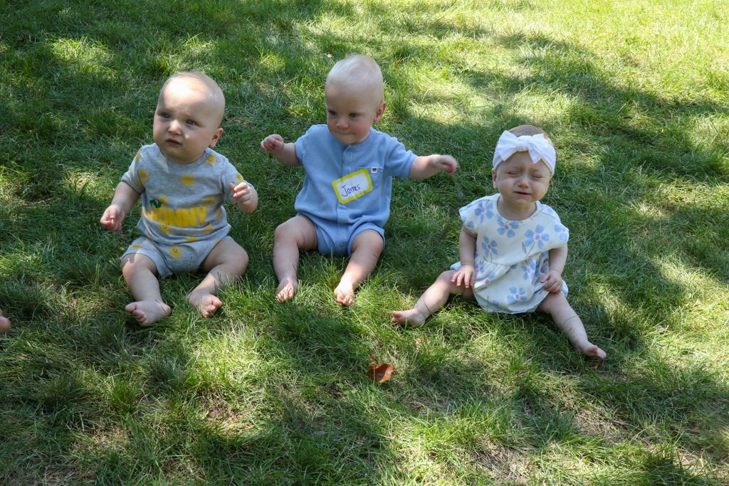 Three babies sitting on grass, two looking up, one with a headband, all in onesies.