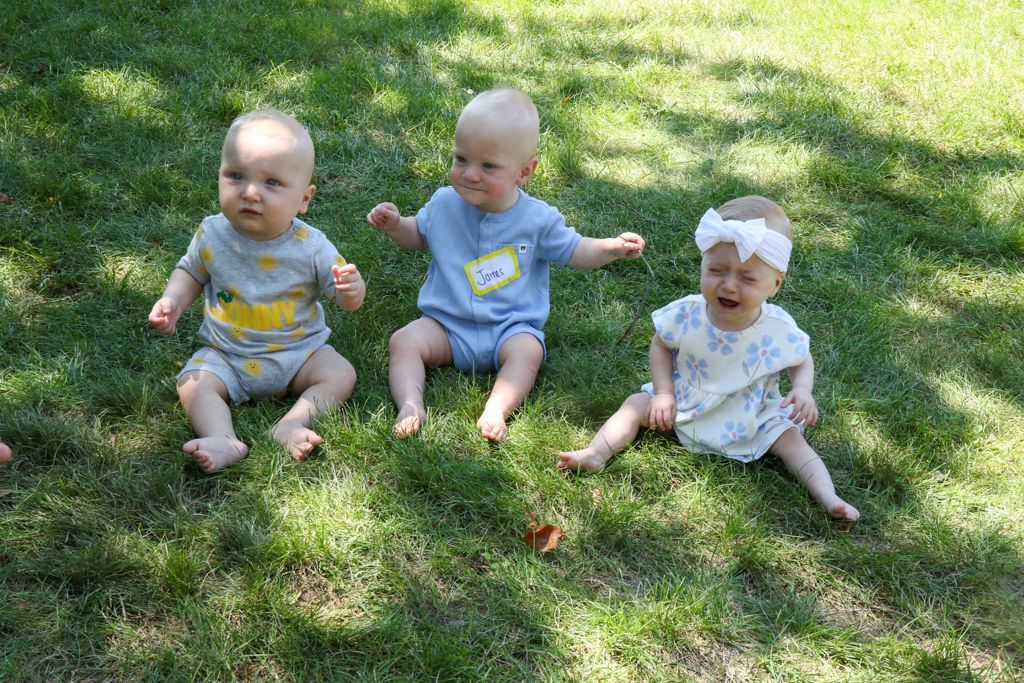 Three babies sit in grass: two calm, one crying, all wearing onesies.
