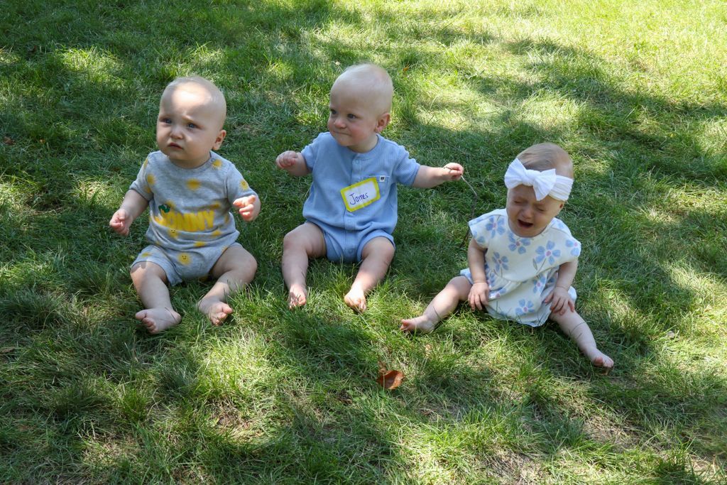 Three babies sitting on grass; two looking forward, one crying with white headband.