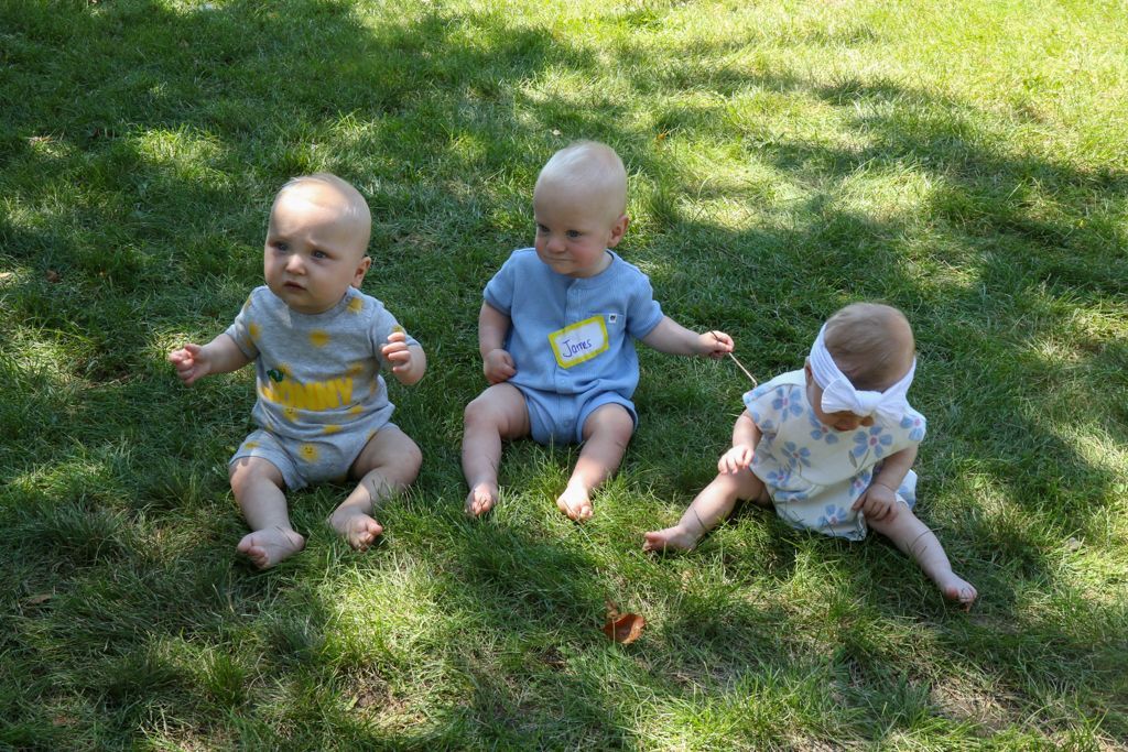 Three babies sitting on green grass, looking up. One has a headband. Sunny day.