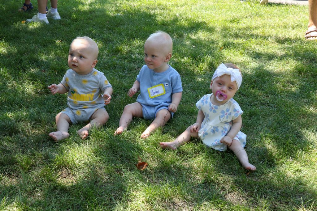 Three bald babies sitting on grass, looking upwards. One has a pacifier and headband.