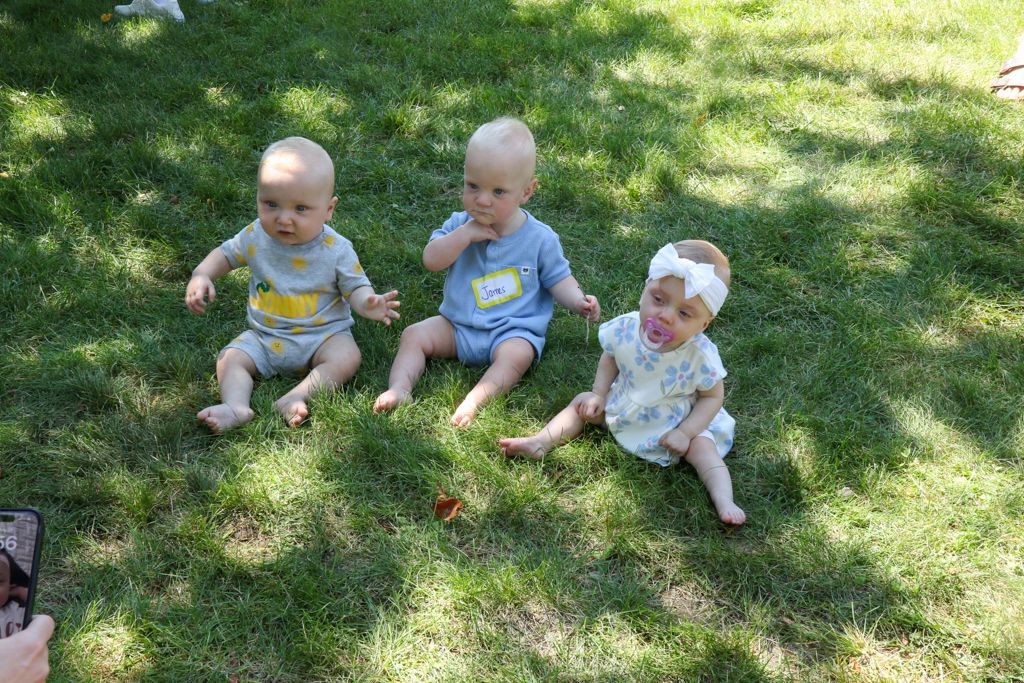 Three babies sitting on grass. One has a pacifier and headband, others in onesies.