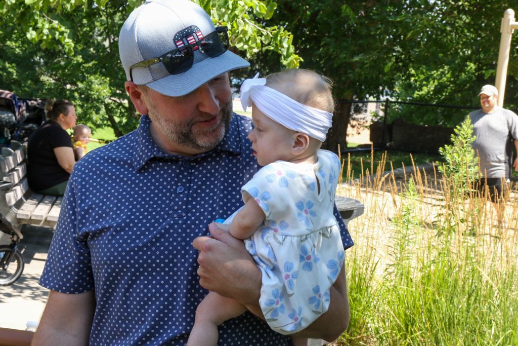 Man holding baby in a park, both looking down. Sunny day, greenery in the background.