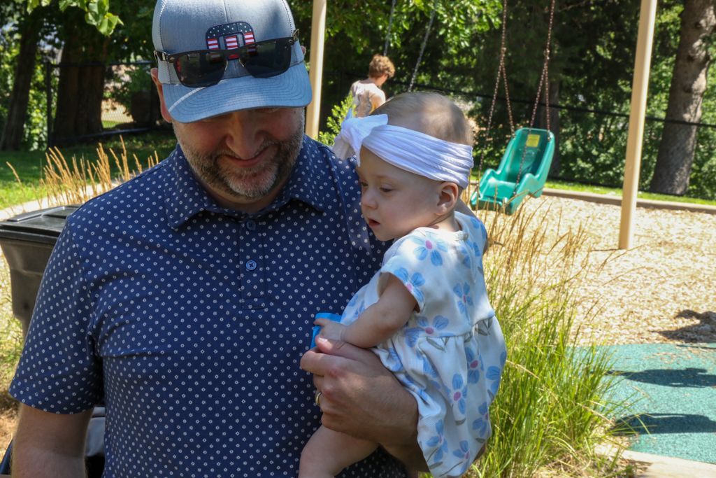 Man in blue shirt holding baby wearing a white bow; playground in background.
