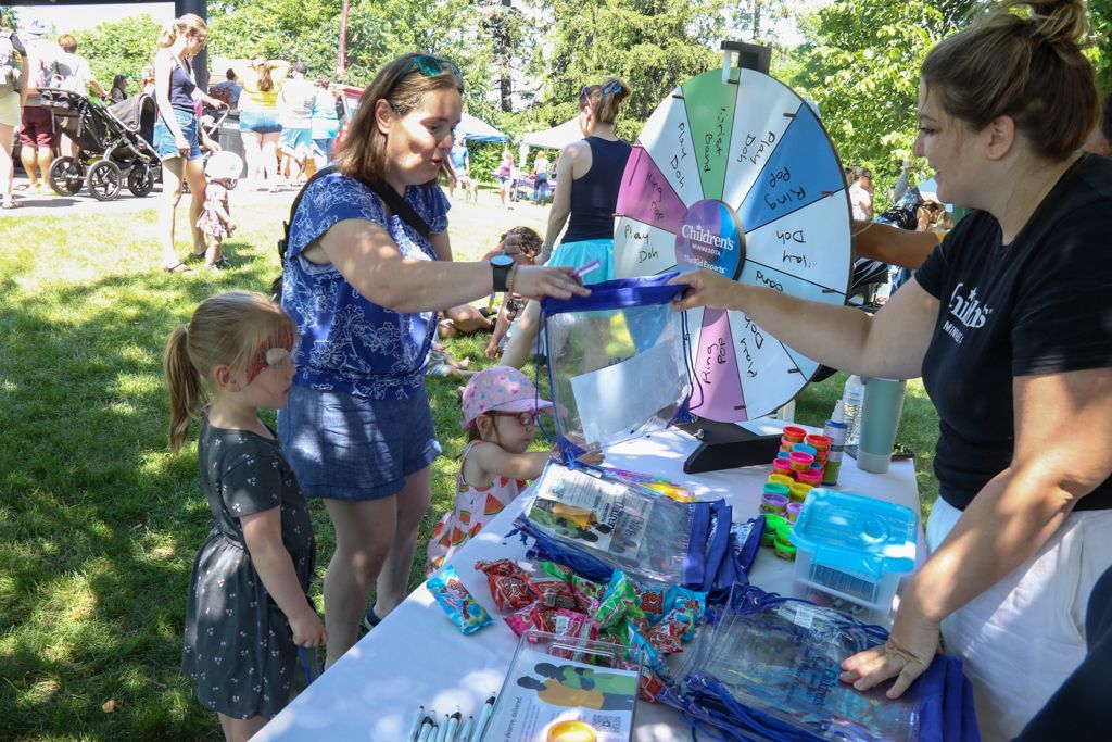 People at a fair interacting with a prize wheel. Woman spins wheel, child watches. Other prizes on table.