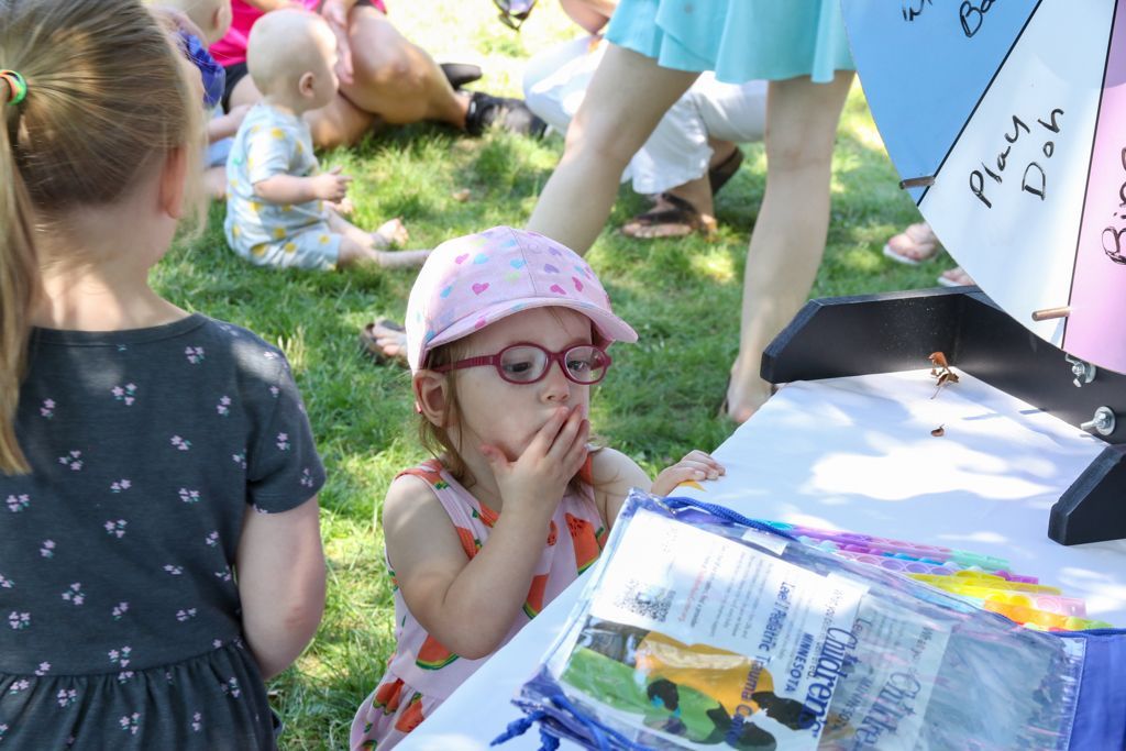 Girl in pink hat with glasses looks at a table with hands near mouth outdoors.