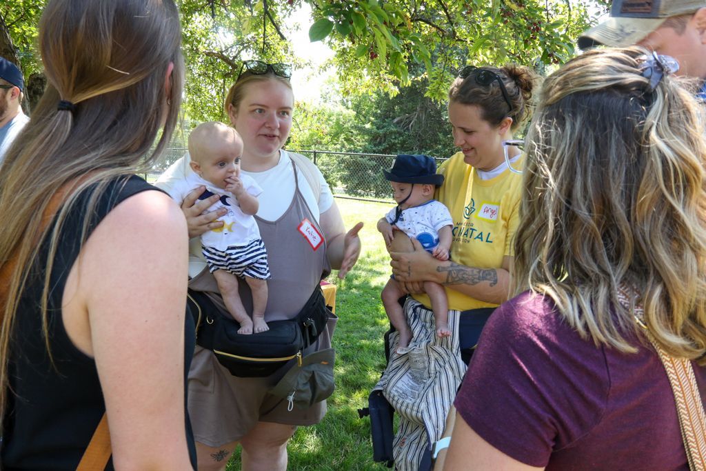 Group of people outdoors, holding babies, conversing under a tree on a sunny day.
