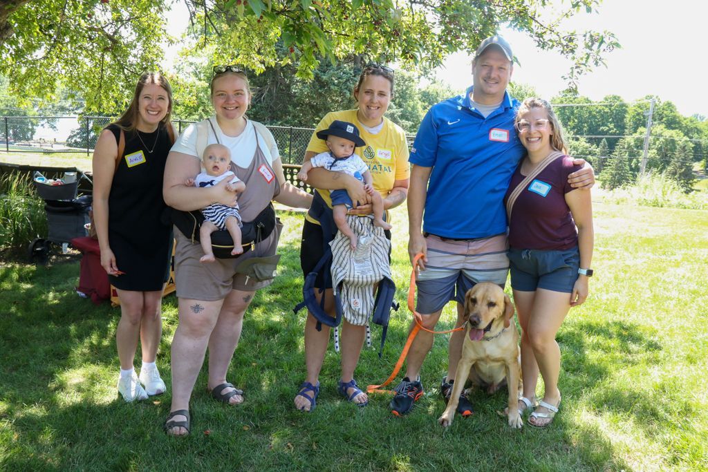 Group of six people and a dog in a park. People are smiling, holding babies, and wearing name tags. Sunny day.
