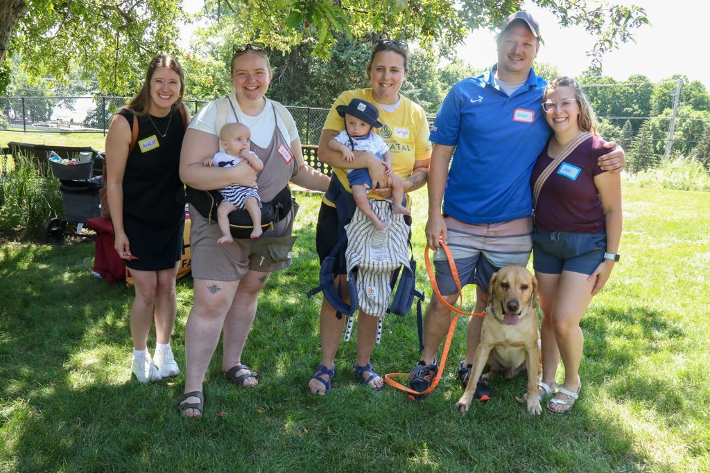 Group of six people and a dog in a park. Two people hold babies. Sunlight.