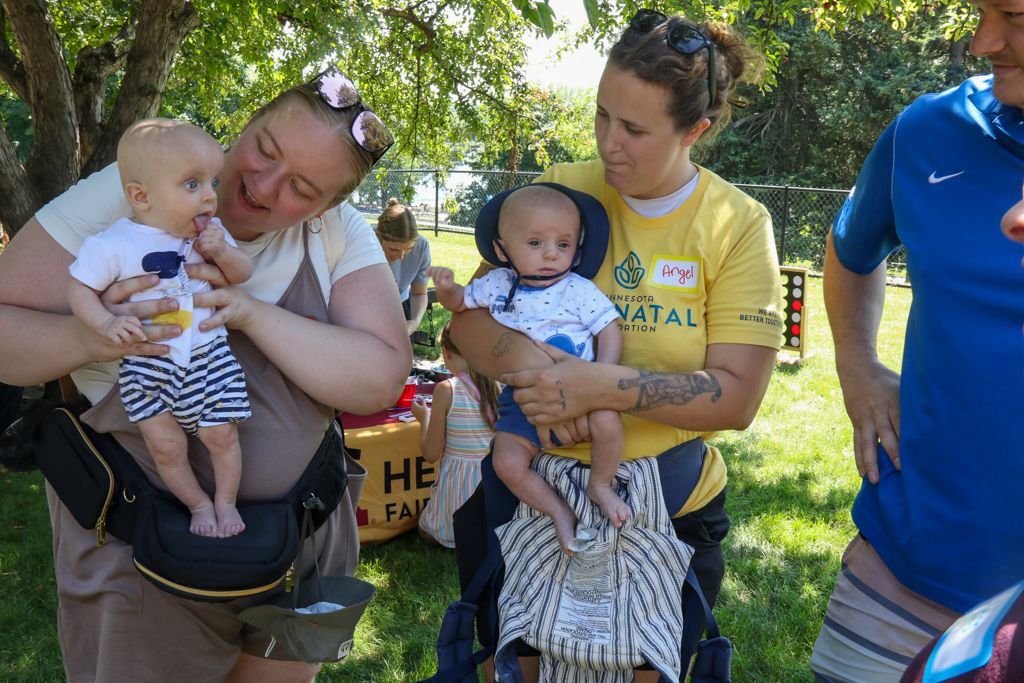 Two women hold babies outdoors; one baby wears a blue shirt, the other a white shirt, all smiling.