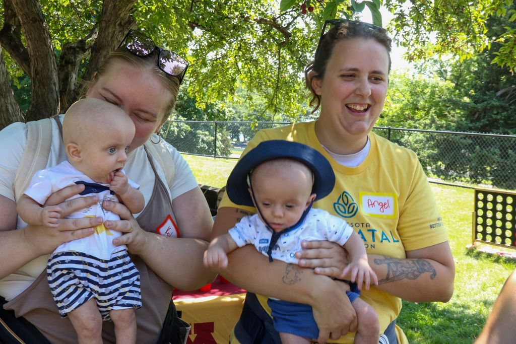 Two women holding babies outdoors, smiling. One baby wears a sun hat. Sunny day.