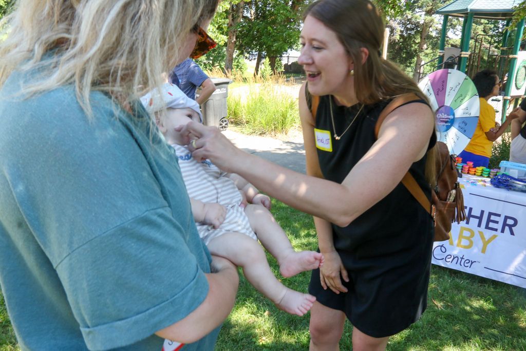 Woman holding baby; another woman smiles, touches the baby's foot outdoors at an event with a prize wheel.