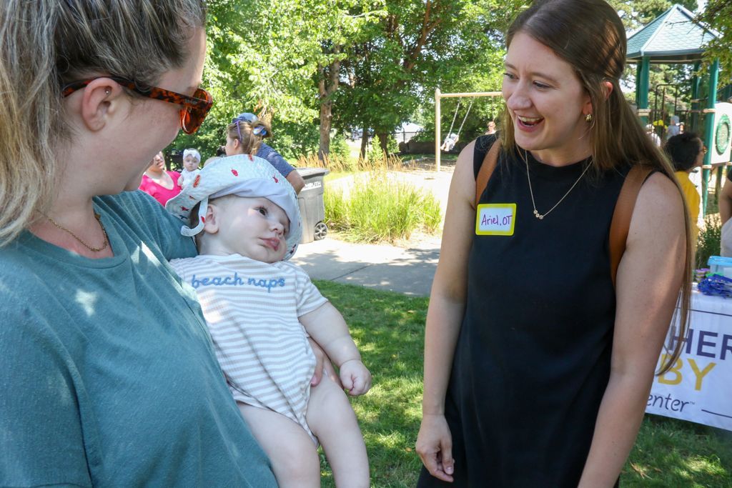 Woman holding baby in park, talking to another woman. Baby wears a hat and 
