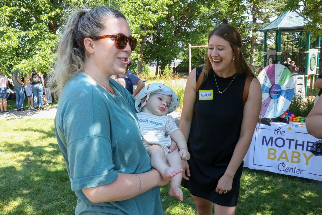 Woman holding baby at outdoor event, smiling; another woman laughs nearby.