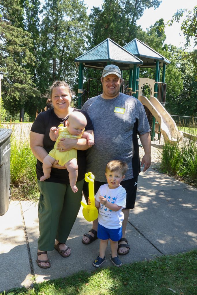 Family of four poses in front of a playground. Mom holds baby, dad stands next to toddler holding a yellow toy.