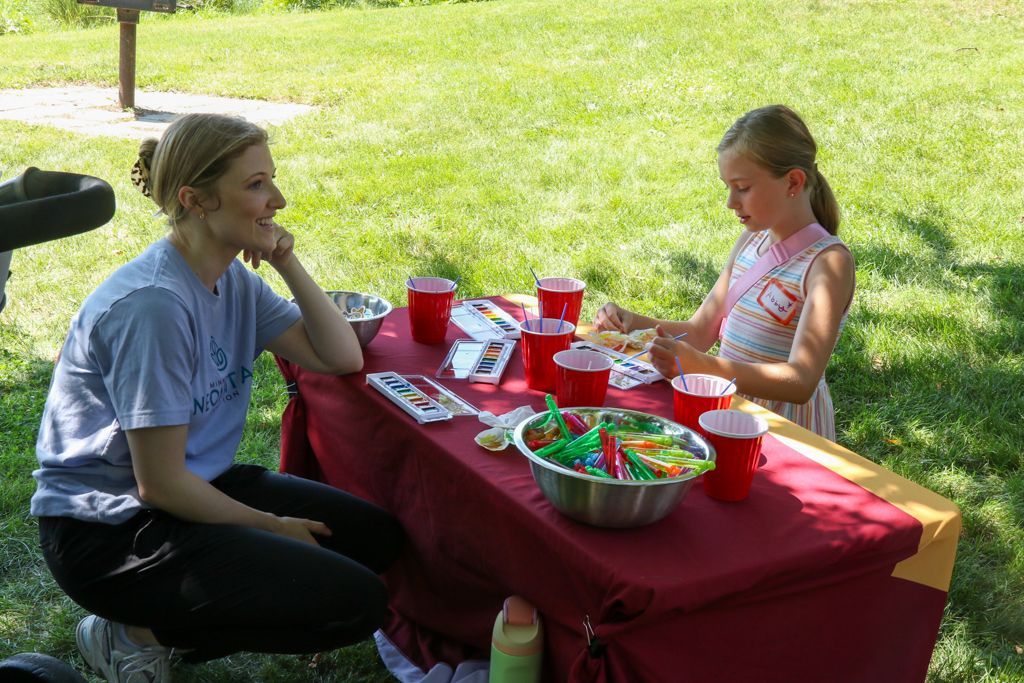 Woman and child at outdoor table with art supplies, drinks. Woman watches child work.