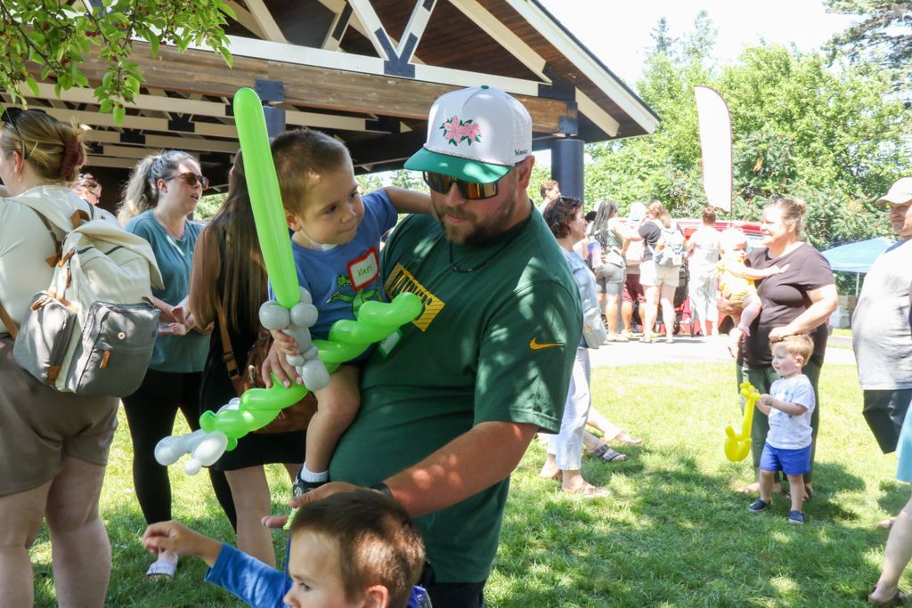 Man holding a child with a green balloon sword in a park with other people.