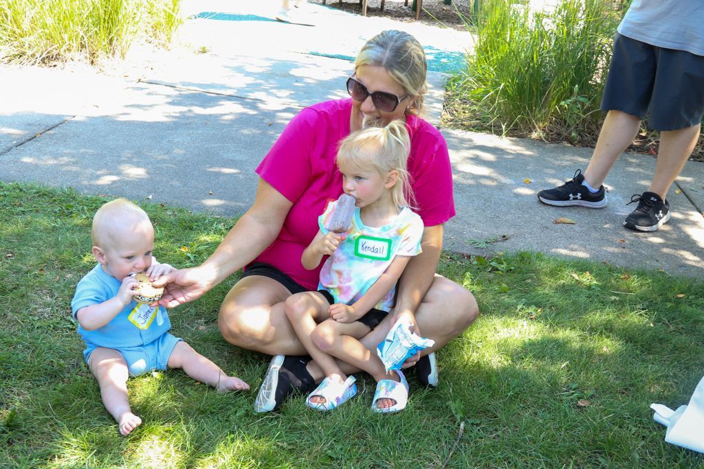Woman sitting on grass with two children, one holding a snack. Outdoors, sunny day.