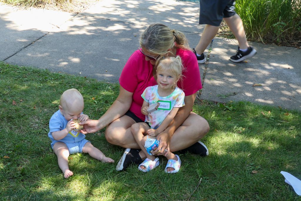 Woman sitting on grass with two children, one holding food, all looking at the camera.