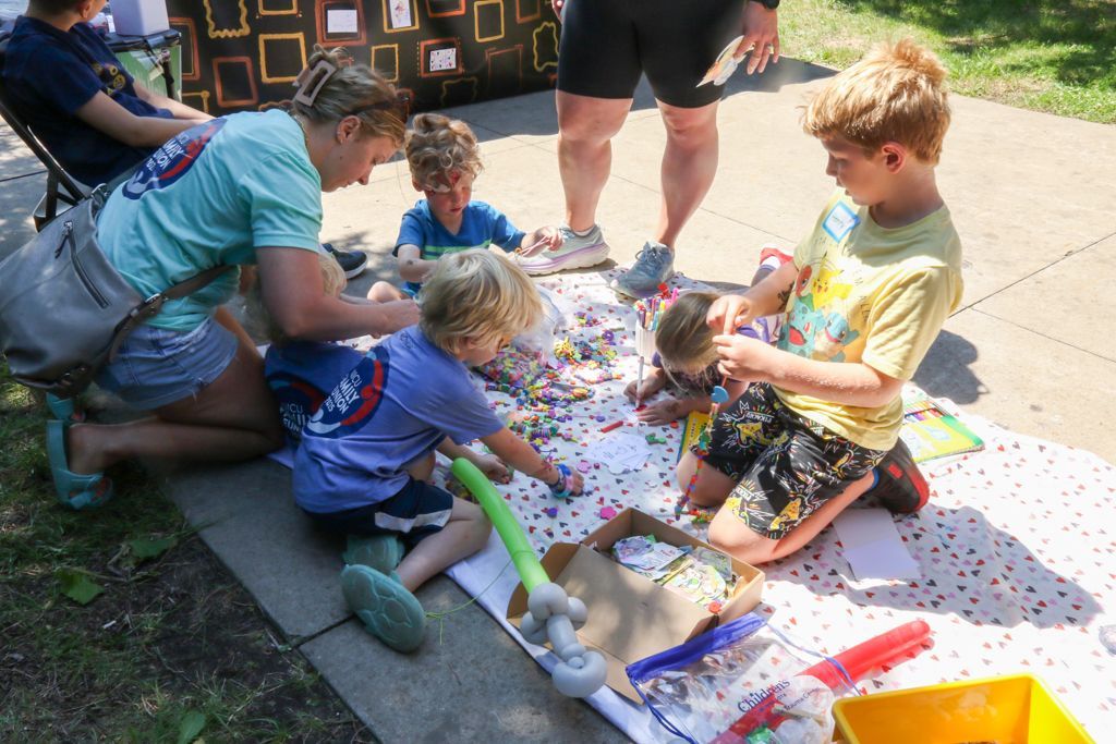 People and children gather on a blanket to sort colorful items outdoors.