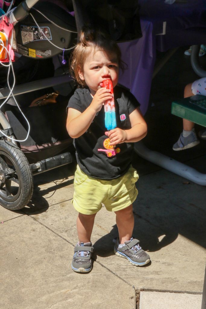 Child eating a popsicle outdoors; wearing a black shirt, green shorts, and gray sneakers.