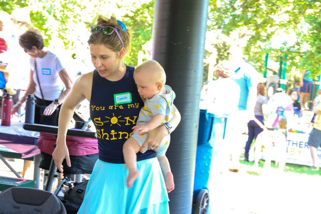 Woman holding baby at outdoor event, wearing blue top and skirt, sunny day.