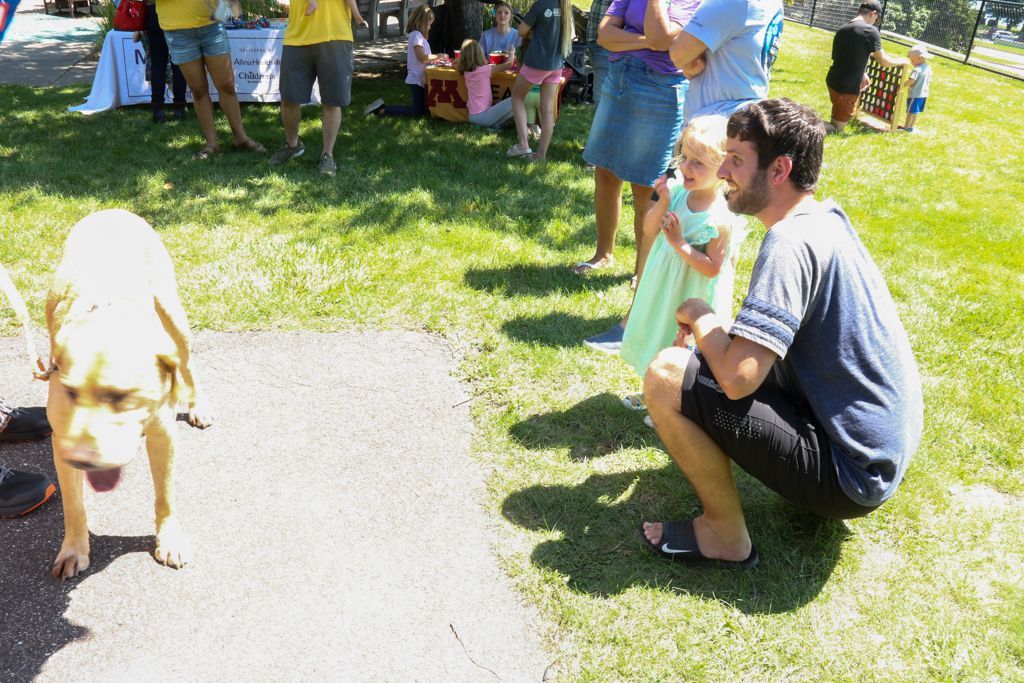 Dog stands on path, looks at man squatting with child on grass at outdoor event.